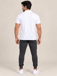 A man modeling a short-sleeve polo shirt in WHITE, paired with black jogger pants and white sneakers, standing against a solid light beige background.