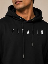 A close-up of a man wearing a black pullover hoodie with the word 'FITAIIM' printed in white across the chest. The hoodie has white drawstrings with silver metal tips and is photographed against a plain beige background.