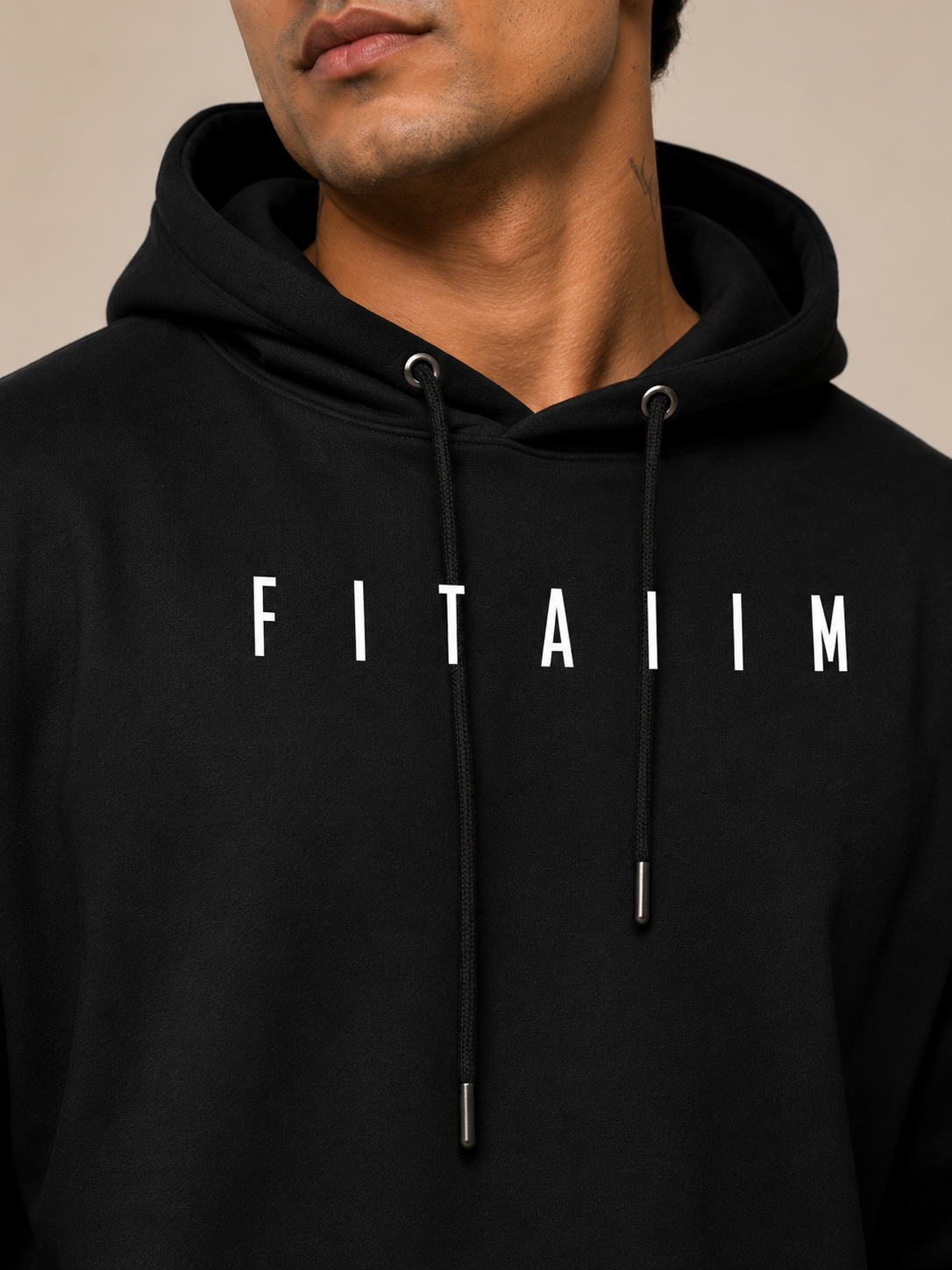 A close-up of a man wearing a black pullover hoodie with the word 'FITAIIM' printed in white across the chest. The hoodie has white drawstrings with silver metal tips and is photographed against a plain beige background.