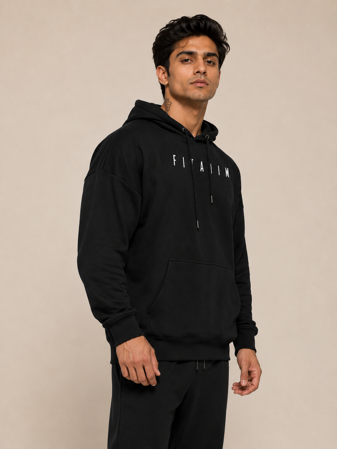 A man wearing a black pullover hoodie with 'FITAIIM' printed in white block letters across the chest, paired with black sweatpants, set against a plain beige studio background.