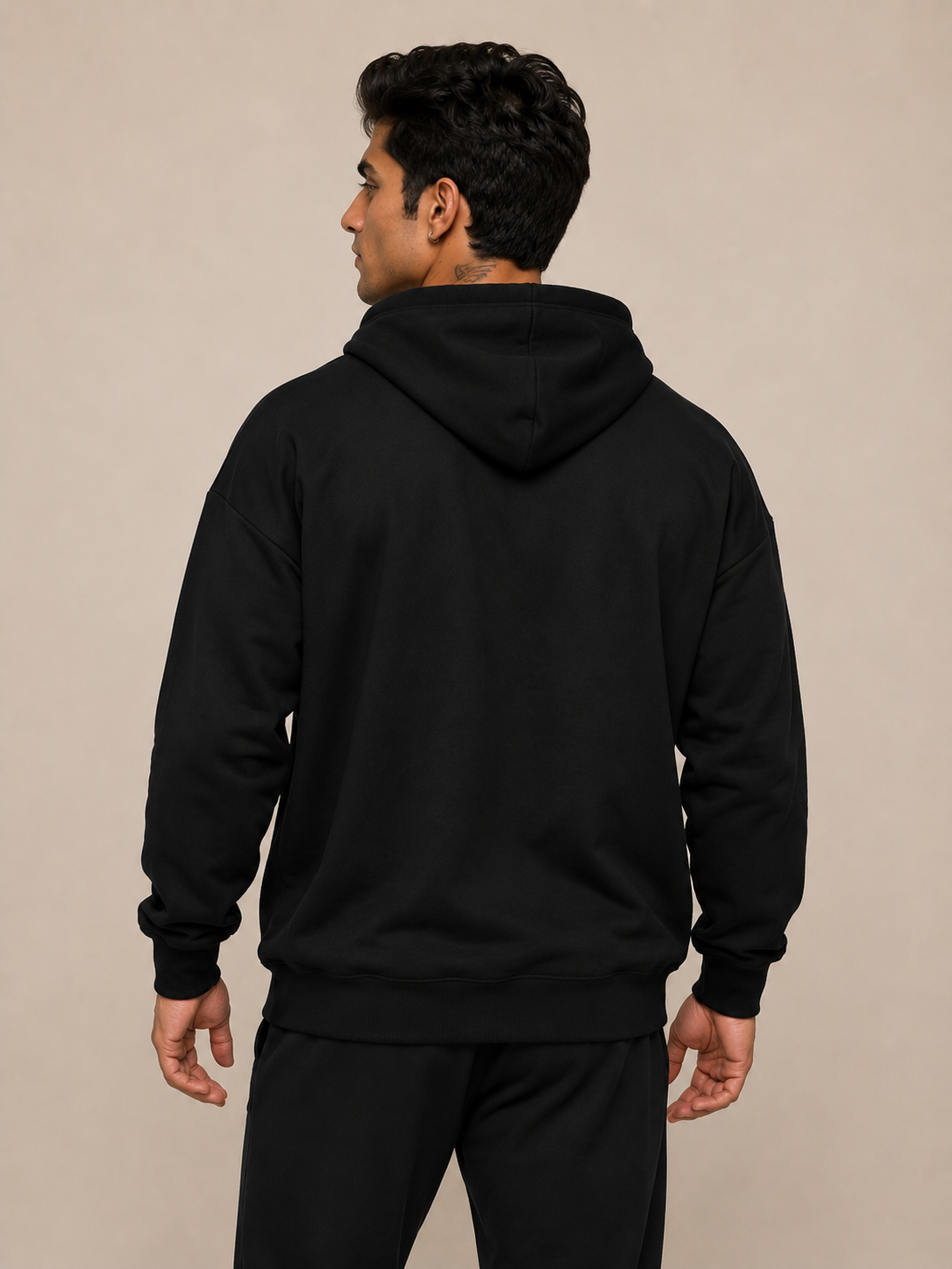 A man wearing a black pullover hoodie and black sweatpants, shown from the back against a plain beige studio background.