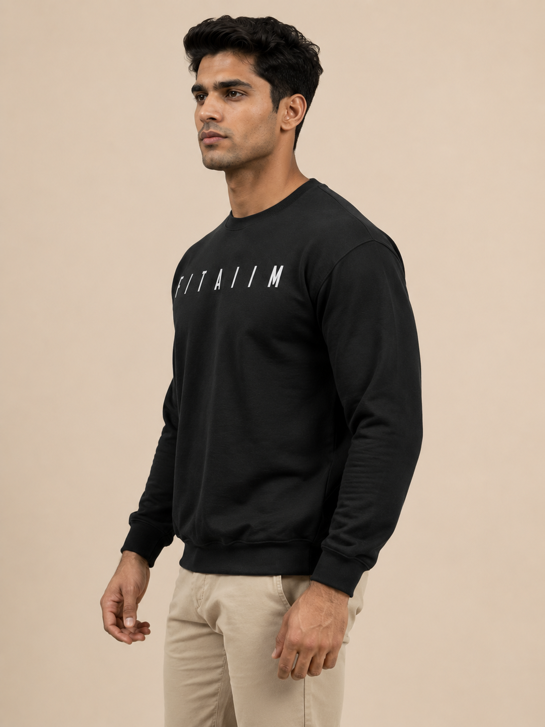 A man wearing a black crewneck sweatshirt with the word 'FITAIIM' printed in white across the chest, paired with beige pants, set against a solid beige background.