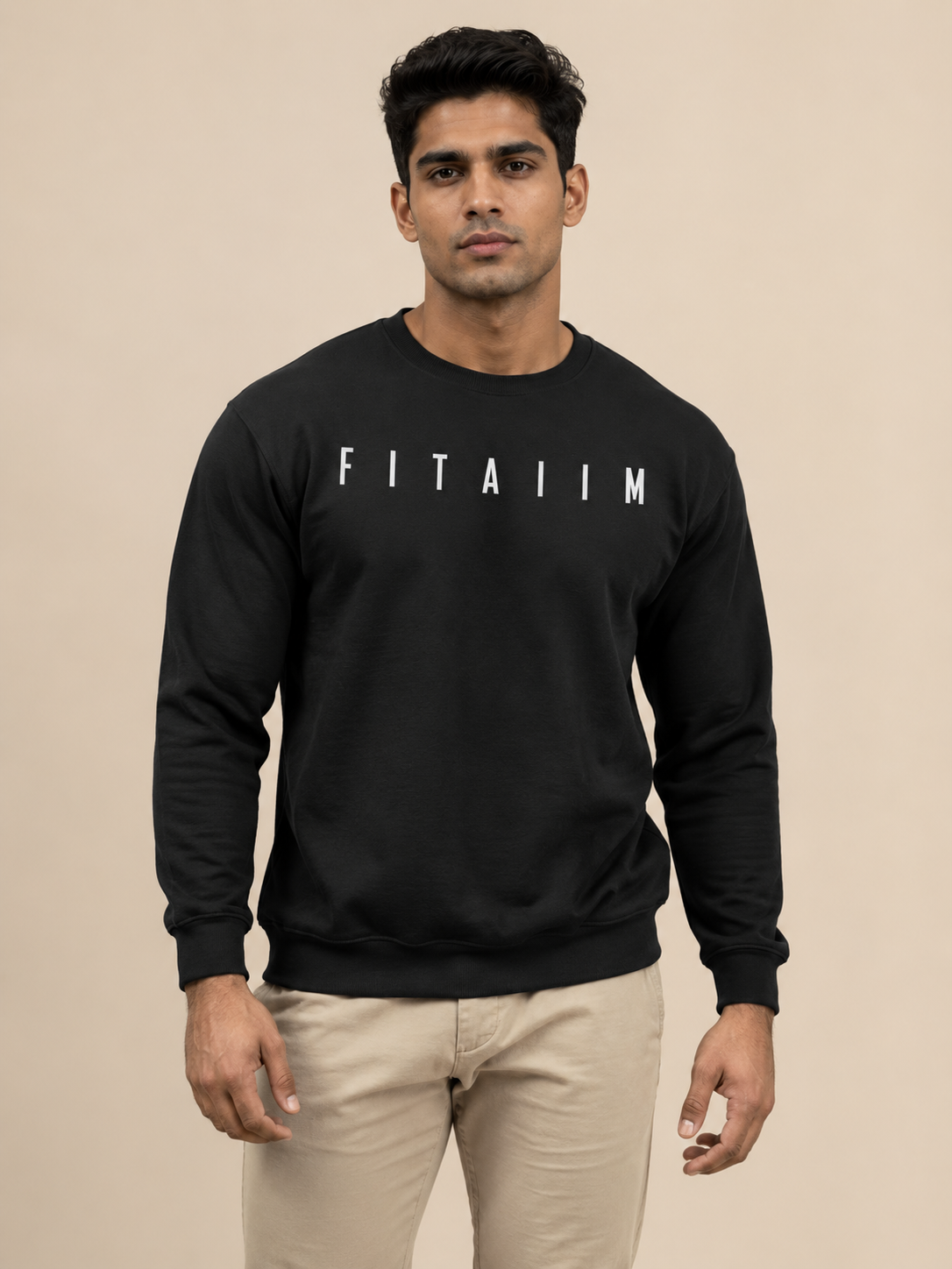 A man wearing a black crewneck sweatshirt with 'FITAIIM' printed in white block letters across the chest, paired with beige trousers, set against a solid beige studio background.