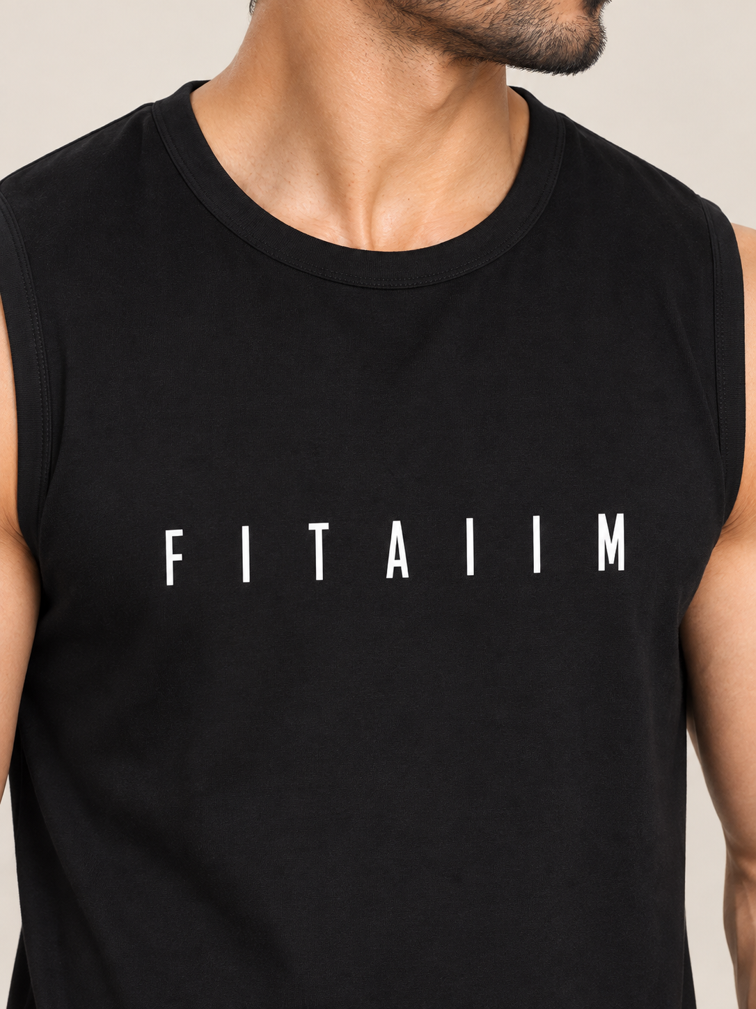 A close-up of a man wearing a black sleeveless muscle tank top with the word 'FITAIIM' printed in white capital letters across the chest, set against a plain beige background.