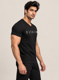 A muscular man wearing a black v-neck t-shirt with 'FITAIWI' printed in white across the chest, paired with white pants, set against a plain beige studio background.