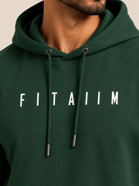 A close-up of a man wearing a dark green hoodie with the name 'FITAIIM' printed in white capital letters across the chest. The shot focuses on the chest area, showing the drawstrings and the text clearly against a neutral beige background.