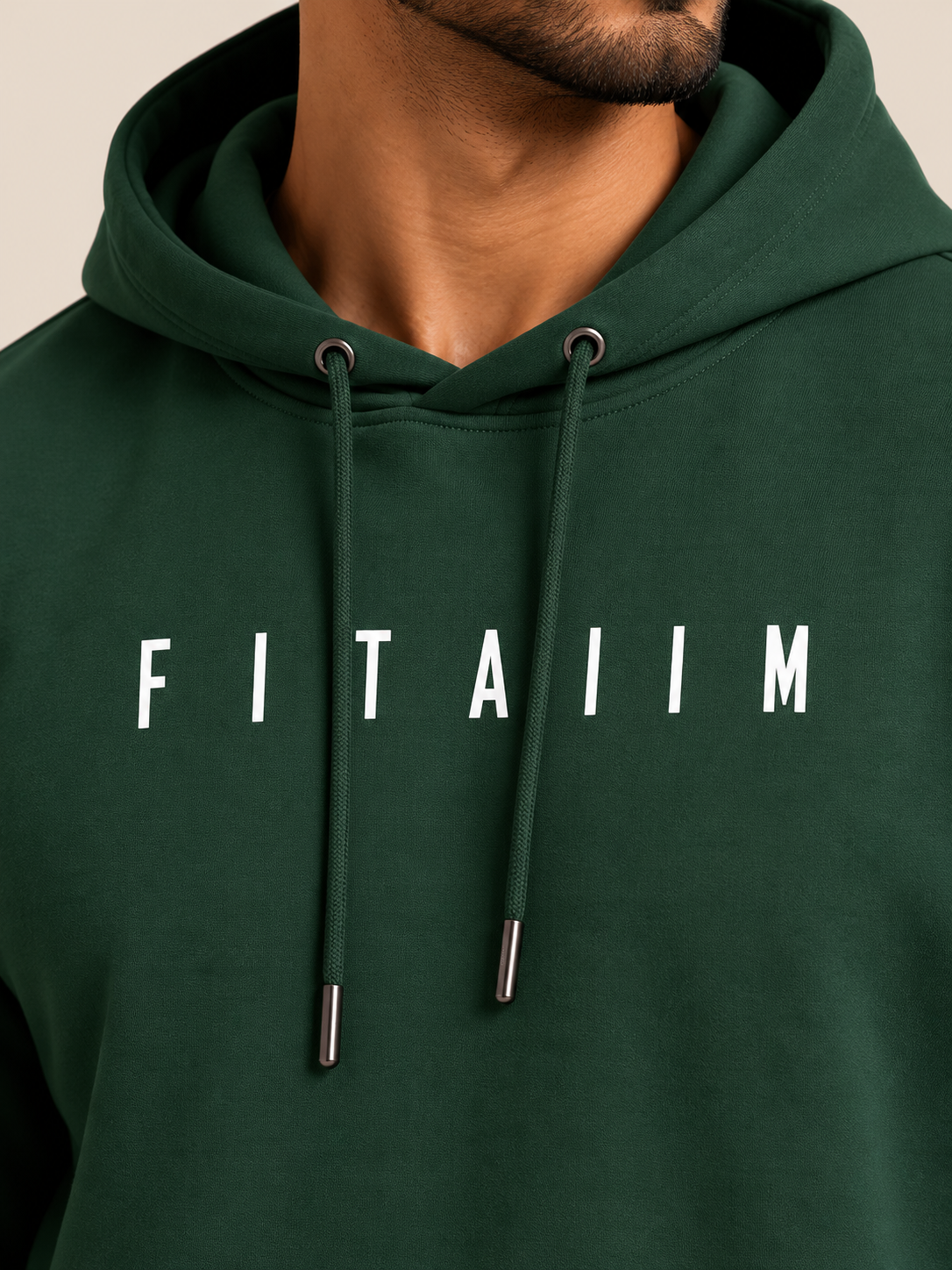 A close-up of a man wearing a dark green hoodie with the name 'FITAIIM' printed in white capital letters across the chest. The shot focuses on the chest area, showing the drawstrings and the text clearly against a neutral beige background.