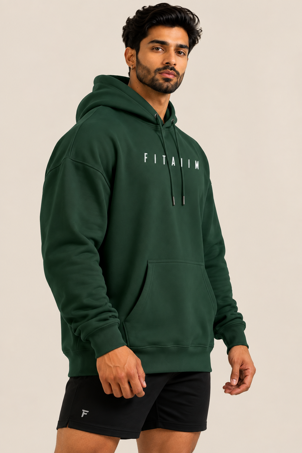 A man wearing a dark green pullover hoodie with 'FITAIIM' printed in white across the chest, paired with black athletic shorts, against a solid light beige background.