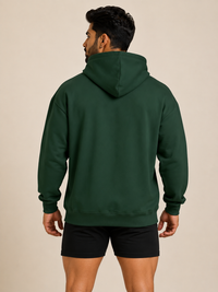 A man shown from the back wearing a dark forest green pullover hoodie and black athletic shorts, standing against a solid beige studio background.