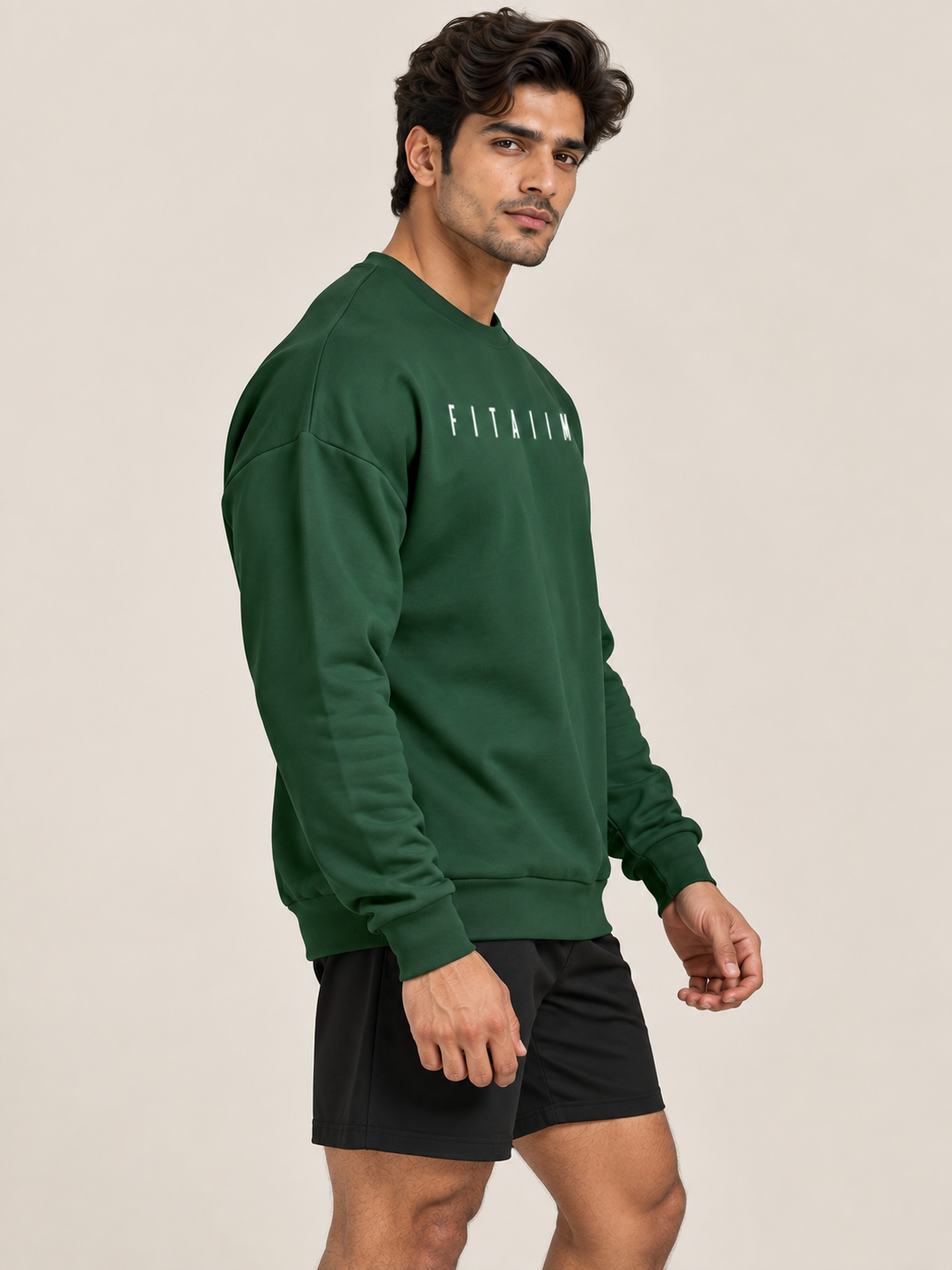 A man modelling a green two-piece tracksuit with white 'FITAIM' text on the sweatshirt and shorts, paired with white sneakers, against a solid beige studio background.