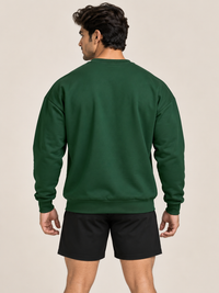 A man viewed from behind wearing a green crewneck sweatshirt and matching black shorts pants, paired with white sneakers, against a plain beige studio background.