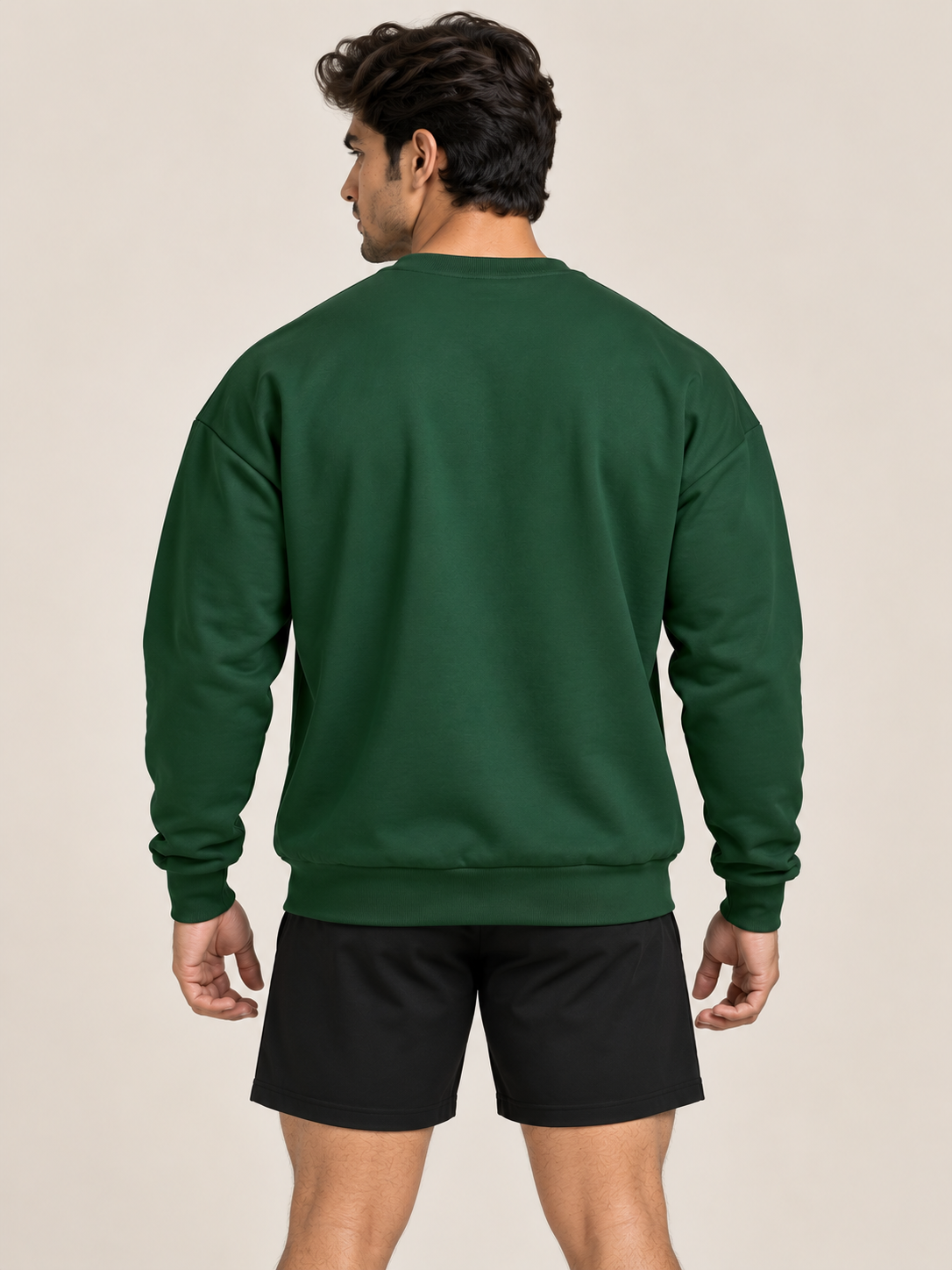 A man viewed from behind wearing a green crewneck sweatshirt and matching black shorts pants, paired with white sneakers, against a plain beige studio background.
