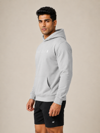 A man wearing a light gray pullover hoodie with a small white logo on the chest, paired with black athletic shorts, standing against a solid beige studio background.