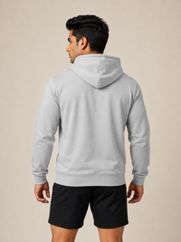 A man shown from the back wearing a light gray pullover hoodie and black athletic shorts, standing against a plain beige studio background.