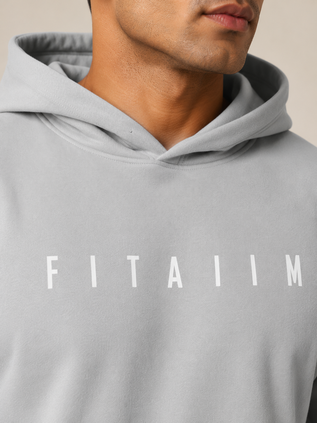 Close-up of a person wearing a light gray pullover hoodie with the word 'FITAIIM' printed in white across the chest, against a plain light beige background.