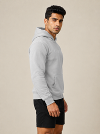 A man wearing a light gray pullover hoodie with a subtle tonal logo on the chest, paired with black athletic shorts, standing against a solid beige studio background.