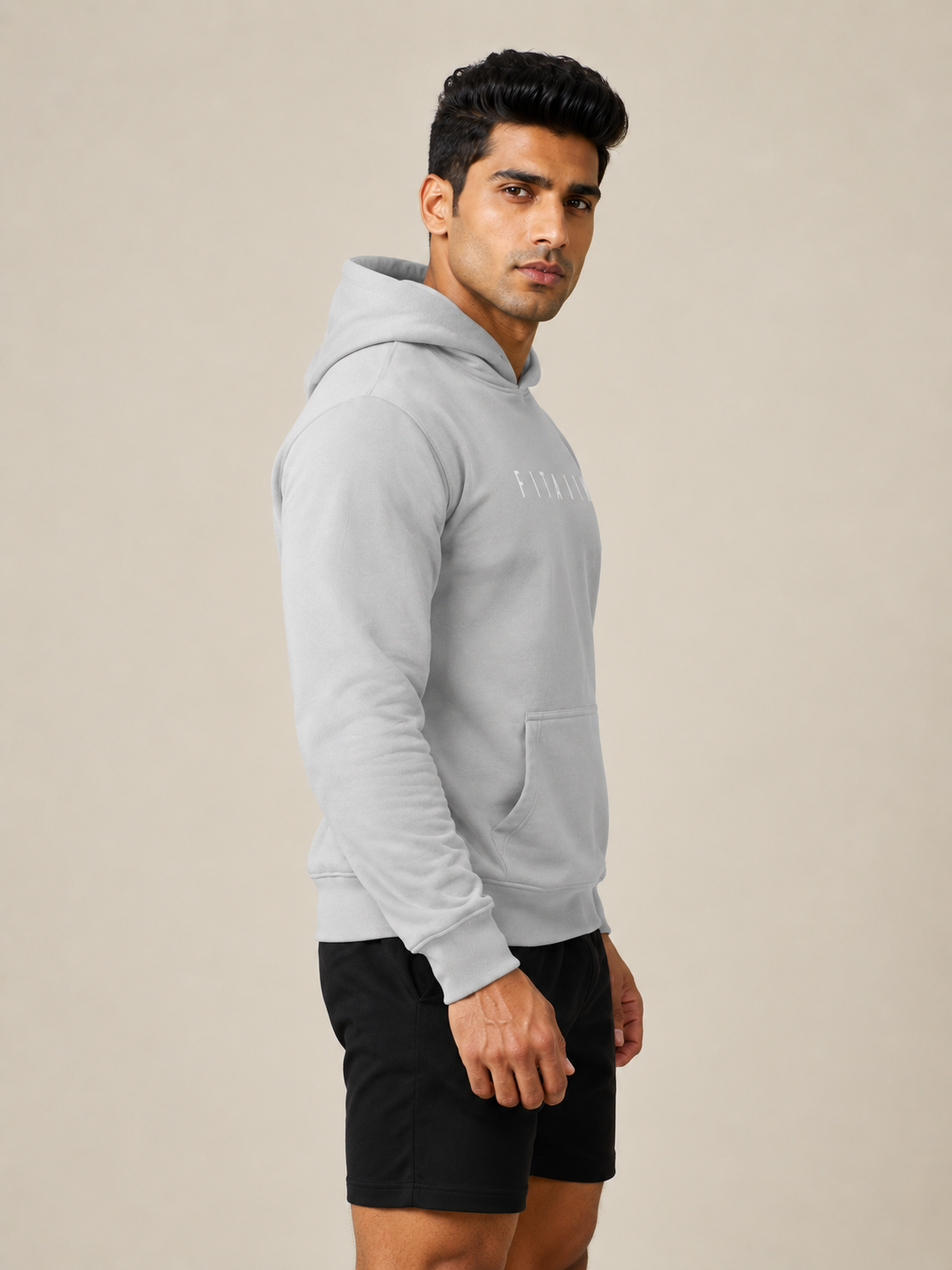 A man wearing a light gray pullover hoodie with a subtle tonal logo on the chest, paired with black athletic shorts, standing against a solid beige studio background.