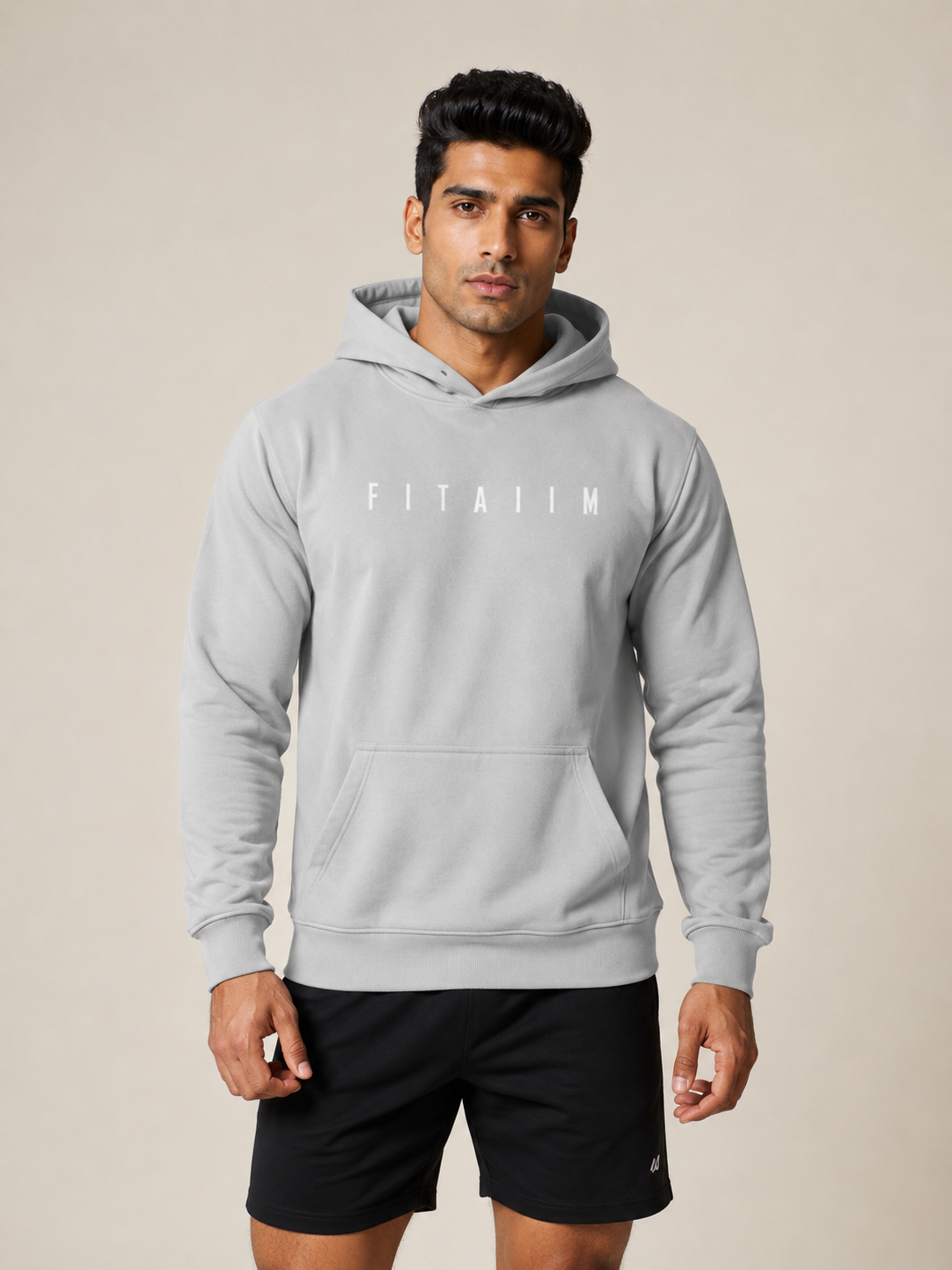 A man wearing a light gray FITA11M pullover hoodie with a white logo on the chest, paired with black athletic shorts, standing against a solid beige background.