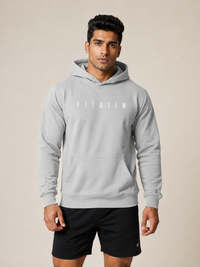 A man wearing a light gray FITA11M pullover hoodie with a white logo on the chest, paired with black athletic shorts, standing against a solid beige background.