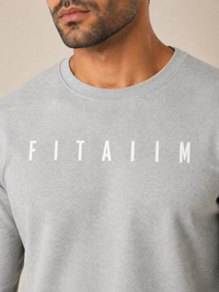 A close-up of a man wearing a light gray crewneck sweatshirt with the word 'FITAIIM' printed in white capital letters across the chest, set against a plain beige background.