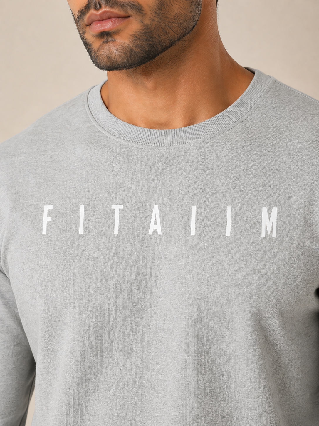 A close-up of a man wearing a light gray crewneck sweatshirt with the word 'FITAIIM' printed in white capital letters across the chest, set against a plain beige background.