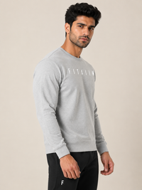 A man wearing a light gray long-sleeve crewneck sweatshirt with 'FITAIIM' printed in white across the chest, paired with black pants, set against a plain beige studio background.
