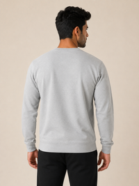 A man viewed from the back wearing a light gray long-sleeve crewneck sweatshirt and black pants, standing against a solid beige studio background.
