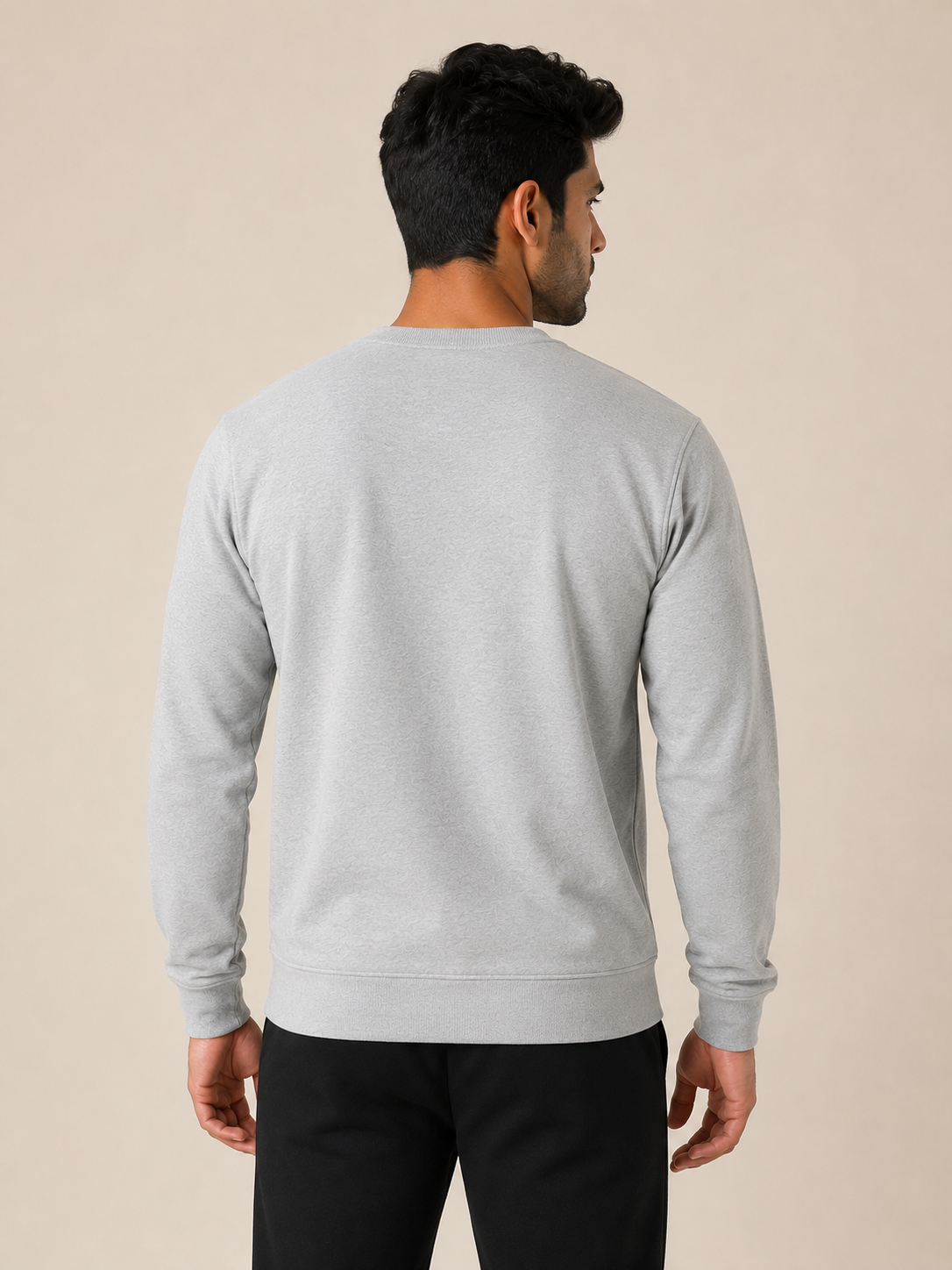 A man viewed from the back wearing a light gray long-sleeve crewneck sweatshirt and black pants, standing against a solid beige studio background.