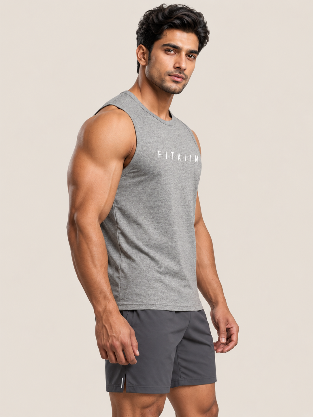 A muscular man wearing a heather gray sleeveless tank top with 'FITAIIM' printed in white across the chest, paired with dark gray athletic shorts, set against a solid light beige background.