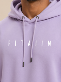 A close-up of a person wearing a light purple/lavender pullover hoodie with white drawstrings and the word 'FITAIIM' printed across the chest in white capital letters.
