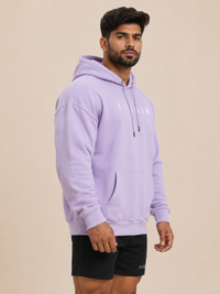 A man wearing a light purple/lavender pullover hoodie with white embroidered text on the chest, paired with black athletic shorts, standing against a solid beige studio background.