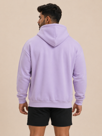 A man viewed from the back wearing a lavender purple pullover hoodie and black athletic shorts, standing against a solid beige studio background.