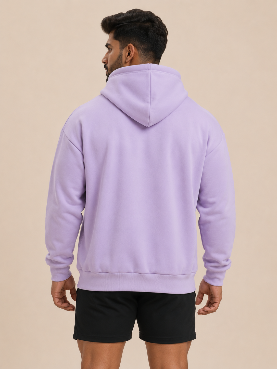 A man viewed from the back wearing a lavender purple pullover hoodie and black athletic shorts, standing against a solid beige studio background.