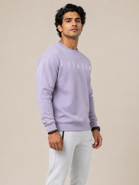 A man wearing a light LAVENDER crewneck sweatshirt with 'FITAIIM' printed in white across the chest, paired with white sweatpants, standing against a solid beige background.