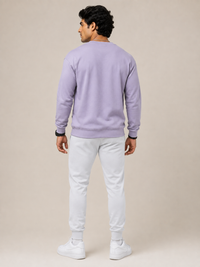A man shown from the back wearing a light LAVENDER crewneck sweatshirt, white jogger pants, and white sneakers, standing against a solid beige studio background.