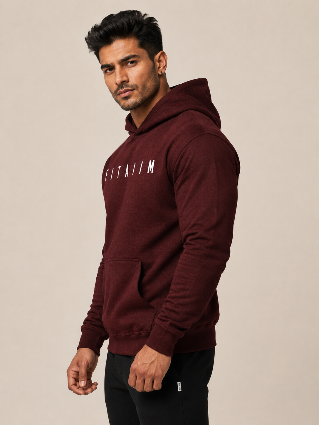 A man wearing a burgundy pullover hoodie with the word 'FITAIM' printed in white across the chest, paired with black pants, photographed against a plain beige studio background.