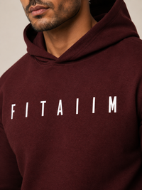 A close-up of a man wearing a maroon hoodie with the word 'FITAIIM' printed in white capital letters across the chest, against a plain light beige background.
