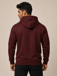 A man viewed from the back wearing a maroon hooded sweatshirt and black pants, standing against a plain beige studio background.