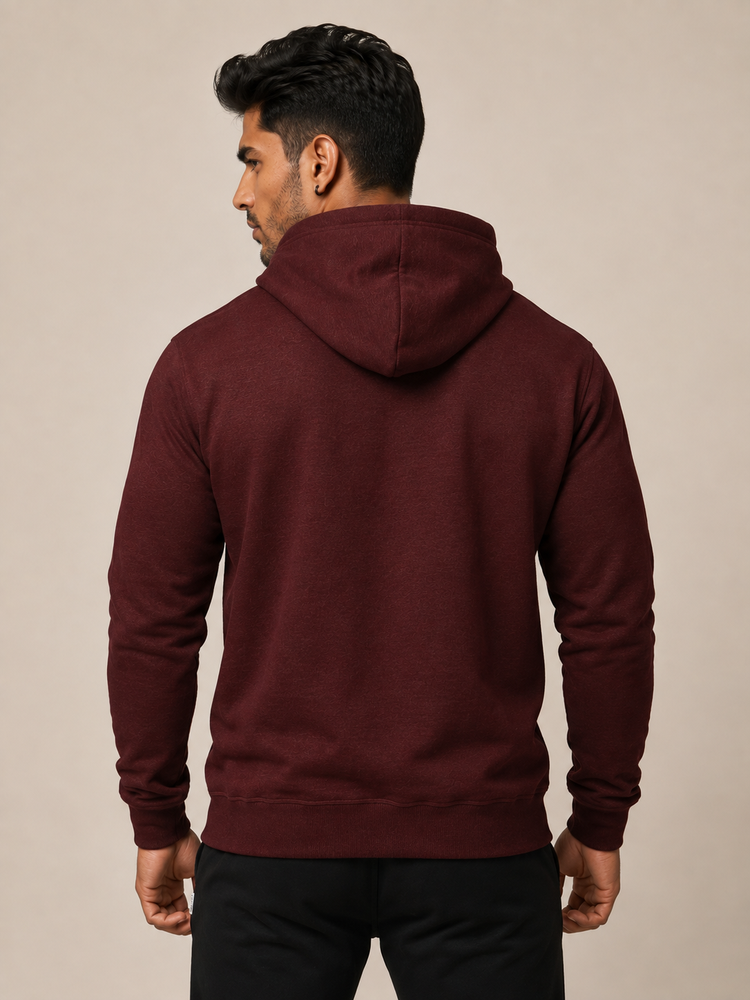 A man viewed from the back wearing a maroon hooded sweatshirt and black pants, standing against a plain beige studio background.