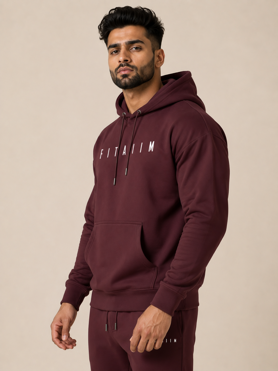 A man wearing a maroon pullover hoodie with 'FITAIM' printed in white across the chest, paired with maroon jogger, photographed against a solid beige studio background.