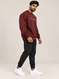 A man modeling a maroon sweatshirt and jogger pants set with white sneakers, standing against a solid beige studio background.