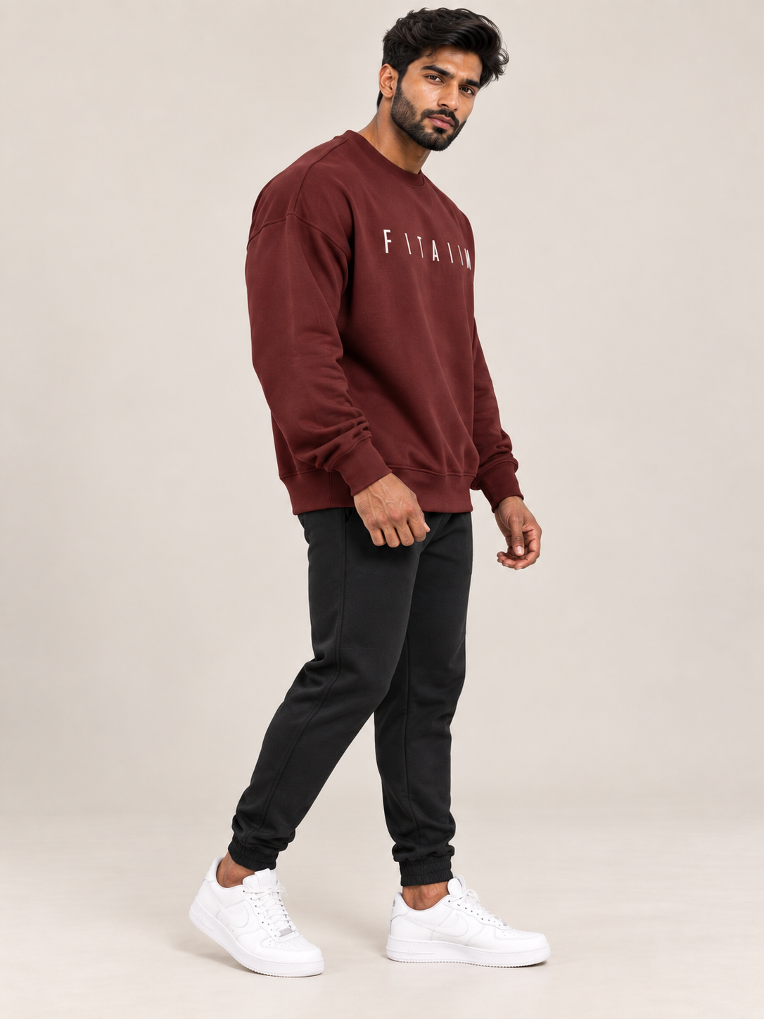 A man modeling a maroon sweatshirt and jogger pants set with white sneakers, standing against a solid beige studio background.