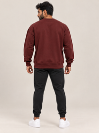 A man viewed from the back wearing a matching maroon sweatshirt and jogger pants set, paired with white sneakers, against a solid light beige studio background.