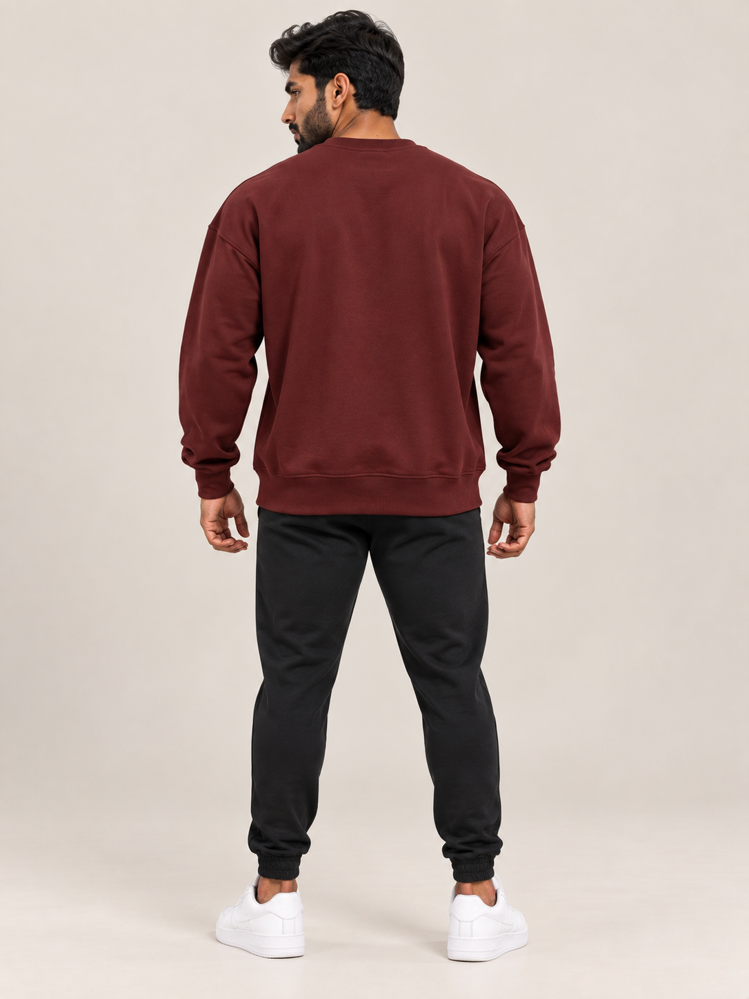 A man viewed from the back wearing a matching maroon sweatshirt and jogger pants set, paired with white sneakers, against a solid light beige studio background.