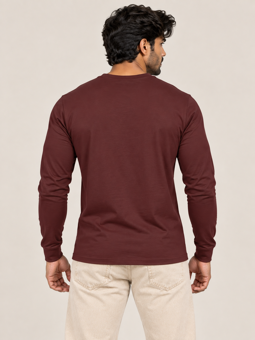 A man viewed from the back wearing a long-sleeve crewneck t-shirt in maroon and beige trousers, against a plain light beige studio background.