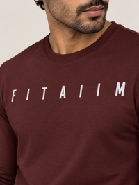 A close-up of a man wearing a maroon long-sleeve t-shirt with the word 'FITAIIM' printed in white across the chest, set against a plain light beige background.