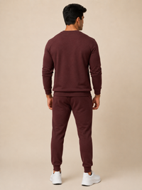 A man viewed from the back wearing a plain maroon crewneck sweatshirt and beige pants, standing against a solid beige studio background.