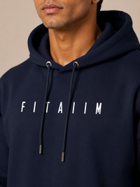 A close-up of a man wearing a navy blue hoodie with the word 'FITAIIM' printed in white across the chest, against a plain beige background.