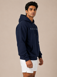 A man wearing a navy blue pullover hoodie with 'FITAIIM' printed in white on the chest, paired with white shorts, standing against a solid beige studio background.