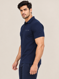 A man modeling a NEAVY BLUE short-sleeve polo shirt with 'FITAIIM' printed on the chest, paired with black joggers and white sneakers, against a solid beige background.